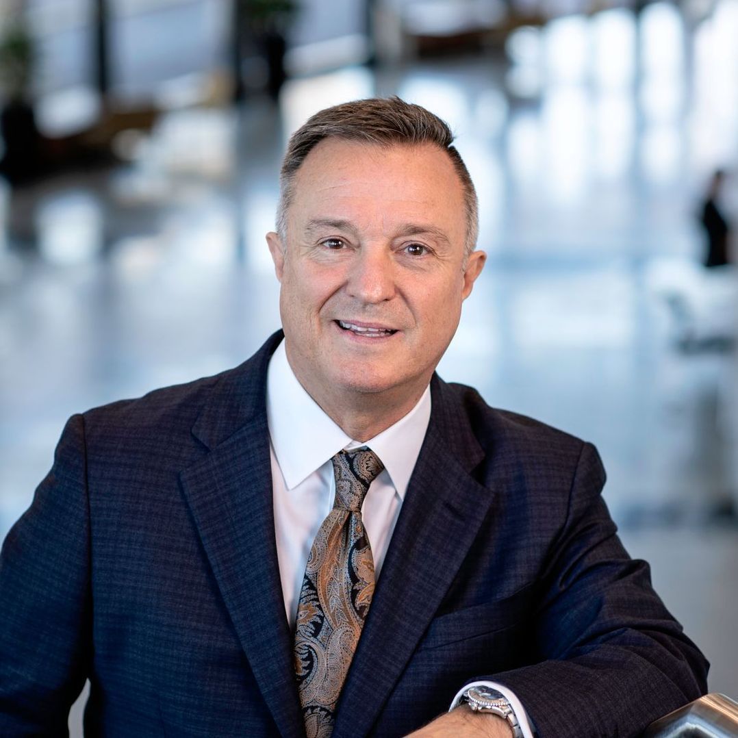 A professional headshot of a person in a dark patterned blazer, white shirt, and patterned tie, smiling in a lobby.