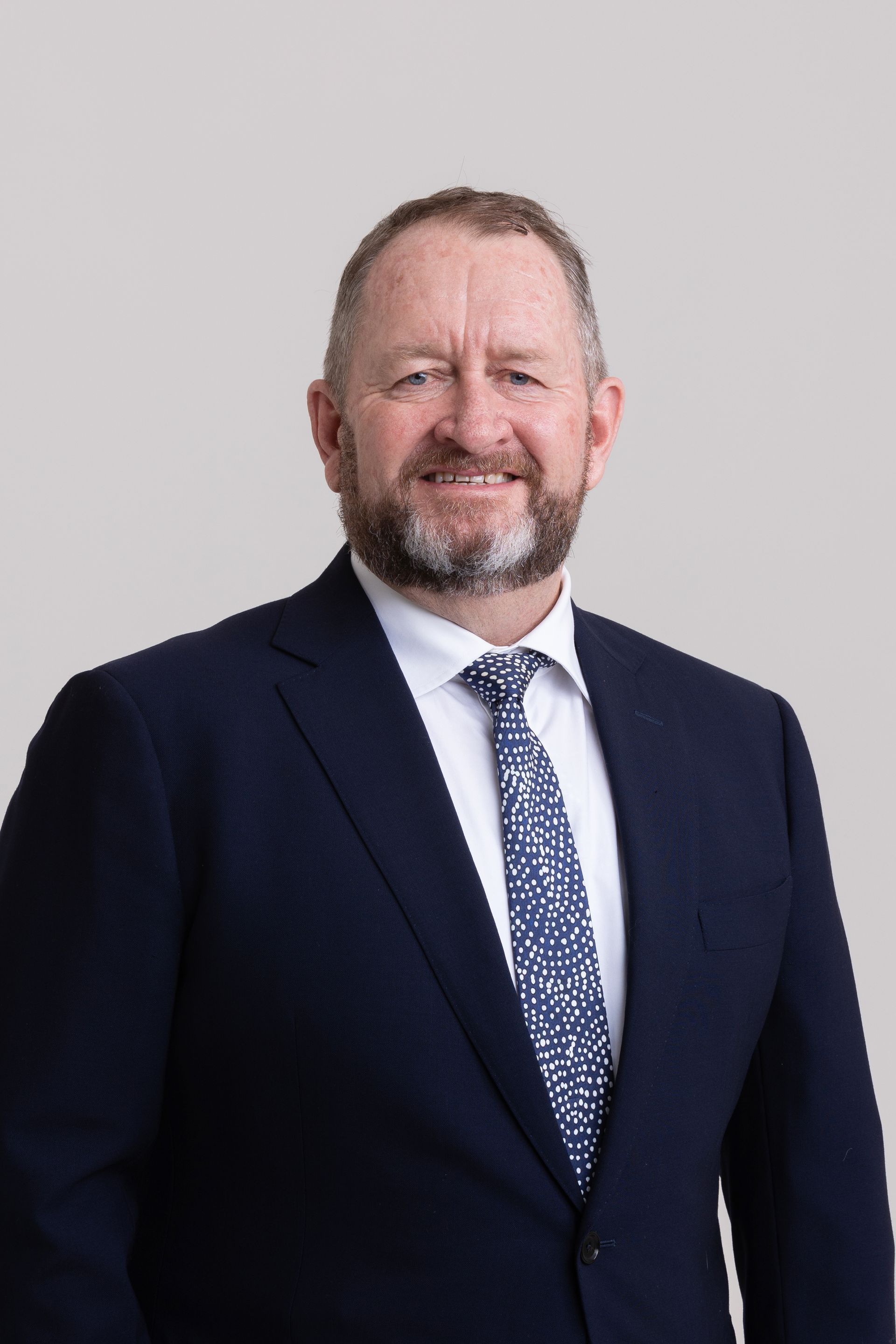 A professional headshot of a smiling person with a beard, wearing a navy blue suit, white shirt, and patterned tie.