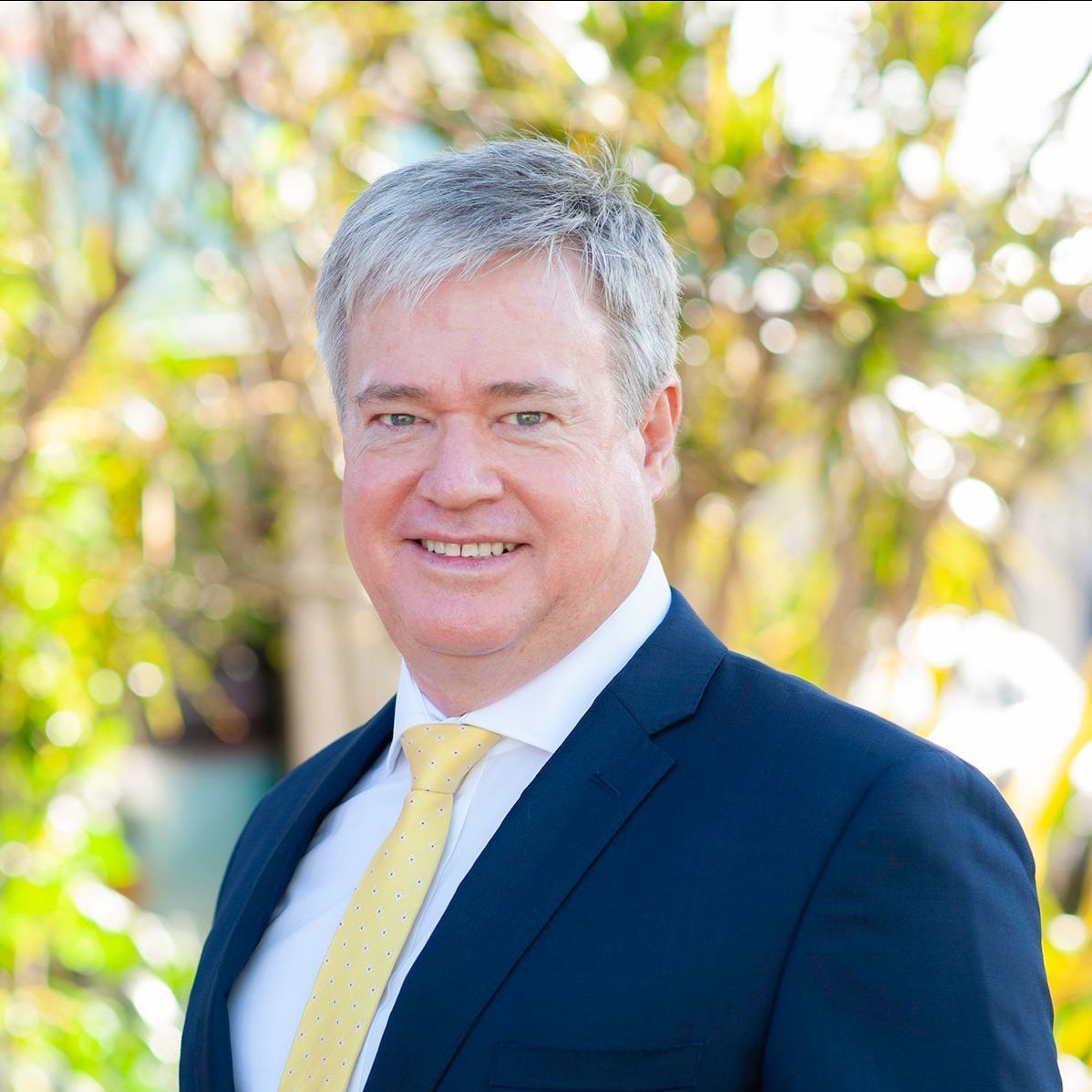 A smiling person in a dark blue suit and a light yellow tie, positioned in front of a blurred outdoor, leafy background.