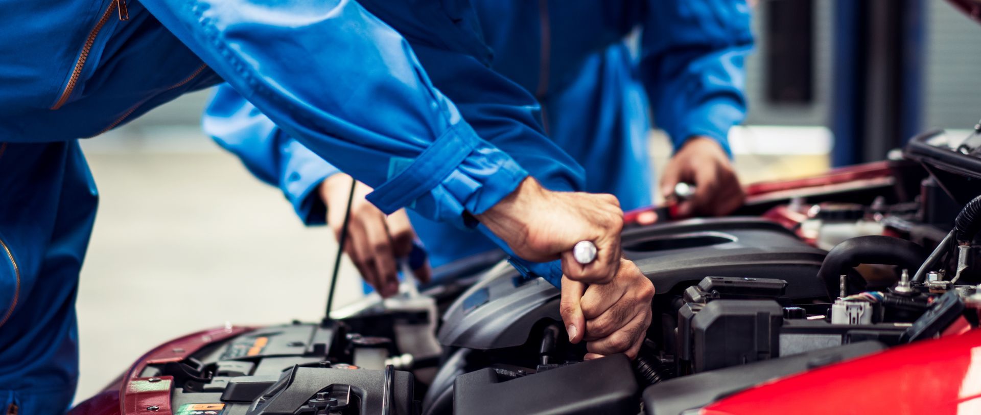 Two mechanics in blue coveralls work on a red car engine.