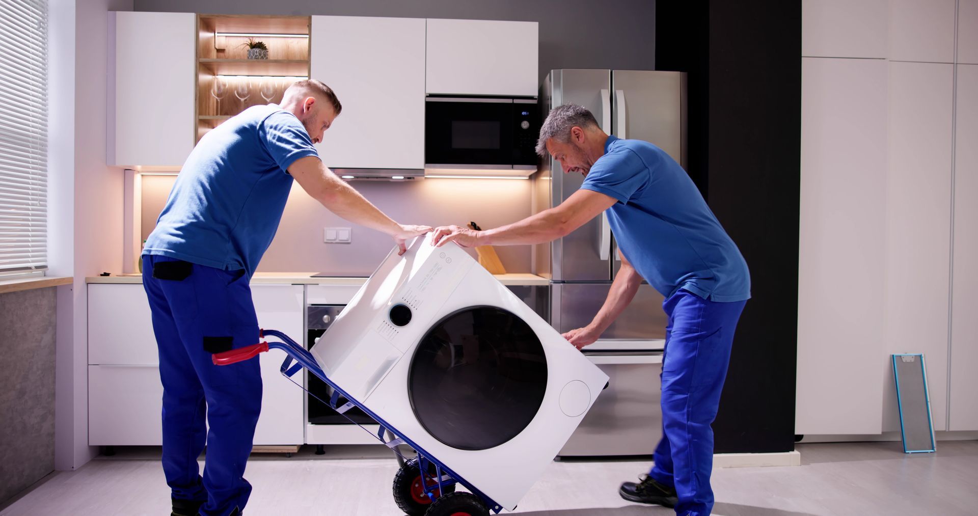 Two workers moving a white appliance on a hand truck in a kitchen.