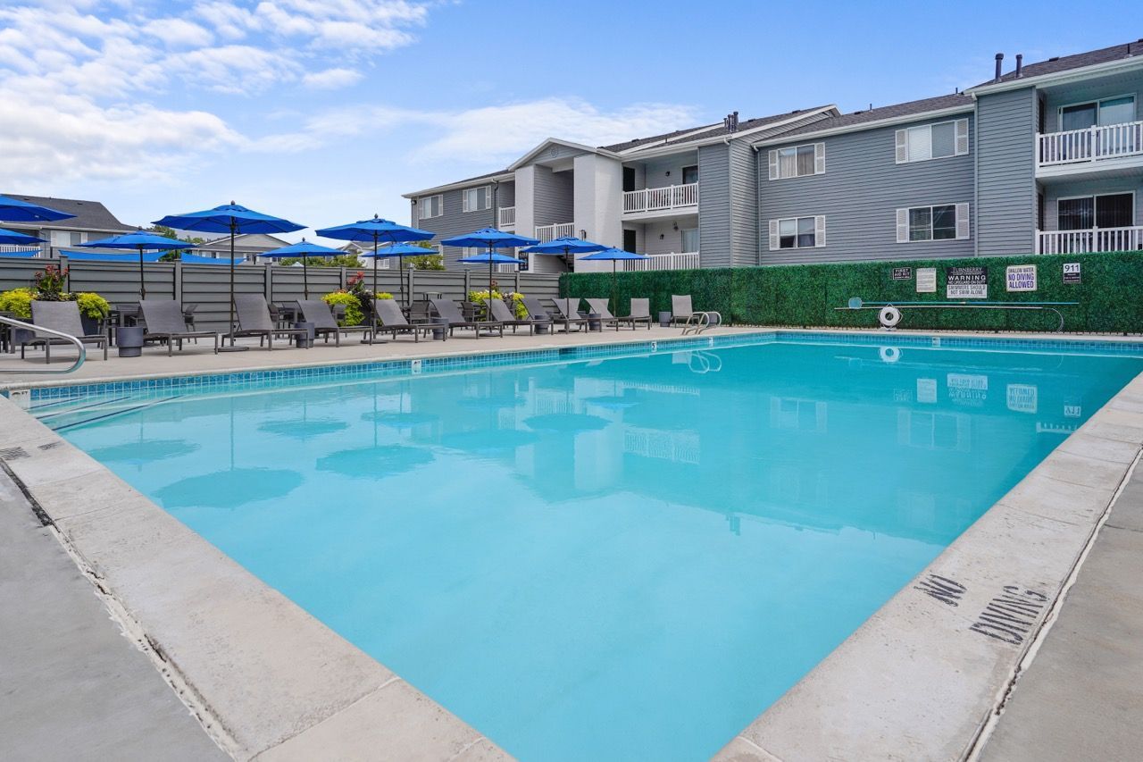 Outdoor pool at an apartment community with blue umbrellas and lounge chairs along a concrete deck. Take a tour of Turnberry Apartments and discover welcoming pet-friendly apartments in Millcreek, UT, where your household can feel at home.