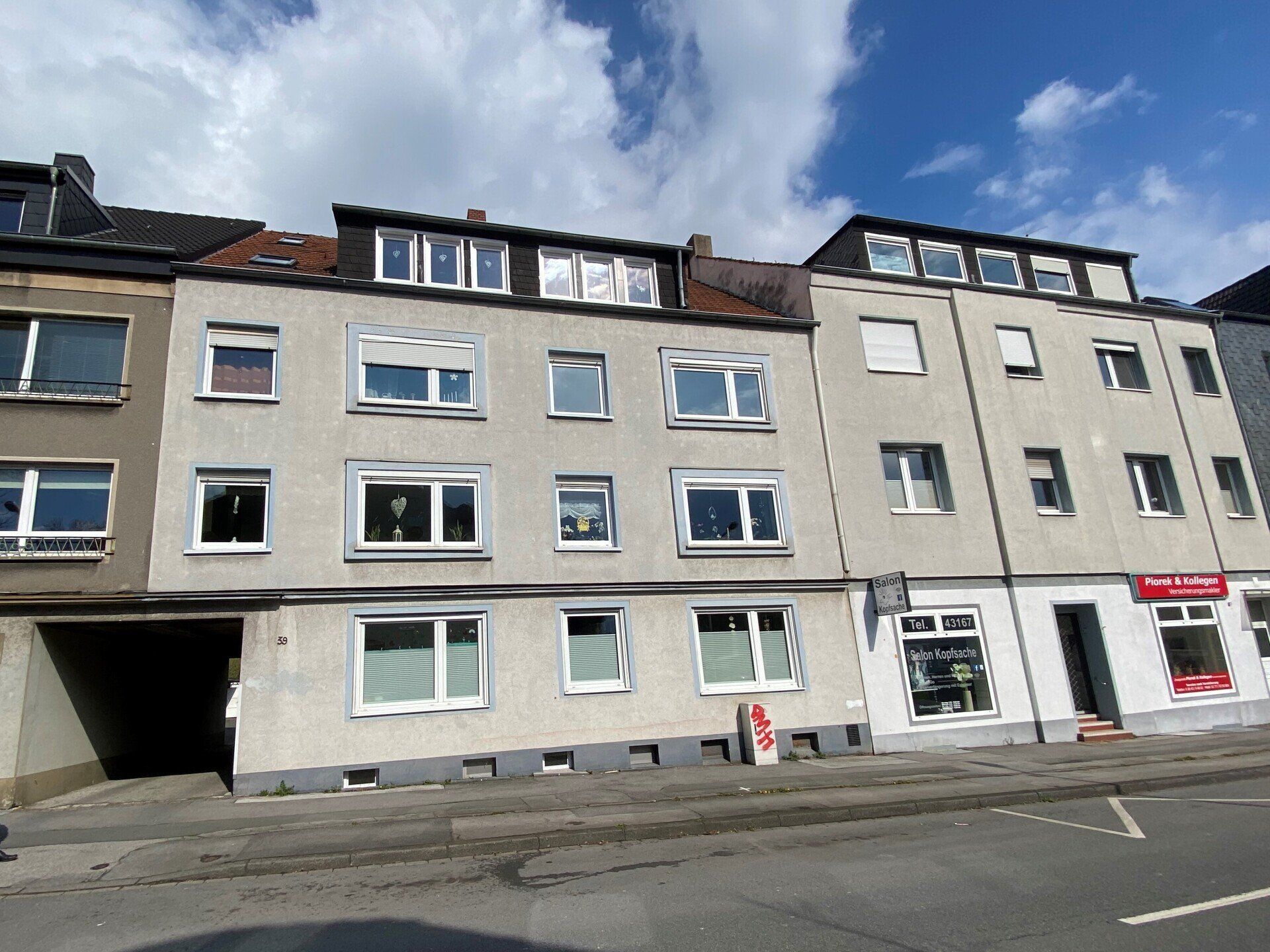 Mehrfamilienhäuser mit mehreren Fenstern, rechts ein Geschäft und im Hintergrund ein bewölkter Himmel.
