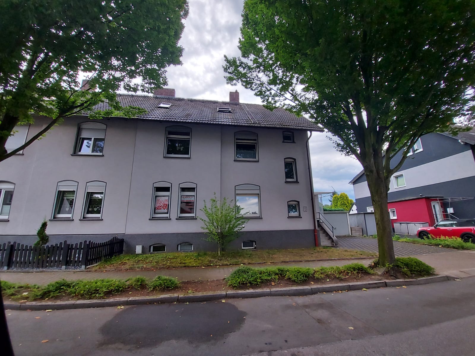 Zweistöckiges graues Gebäude mit mehreren Fenstern, Bäumen an den Seiten und einem kleinen Hof davor.