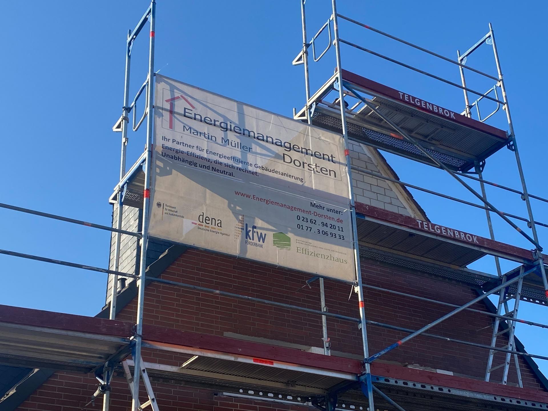 Ein Gerüst an einem Backsteingebäude mit einem Schild mit der Aufschrift „Energie Management Doorten“ vor blauem Himmel.