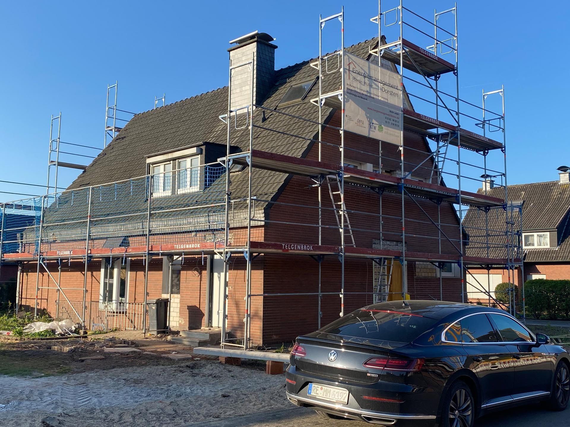 Haus im Bau mit Gerüst, davor parkt ein schwarzes Auto.