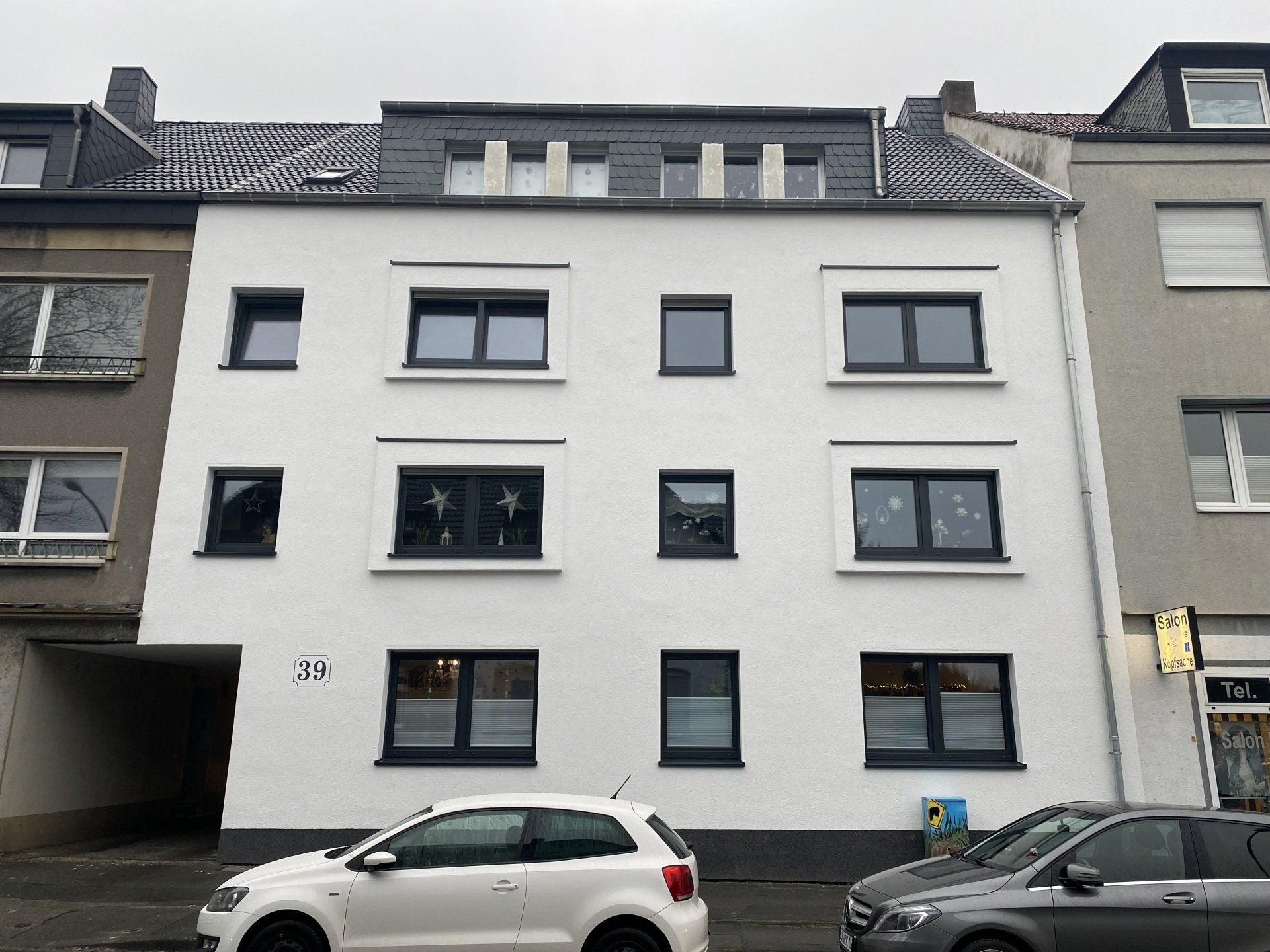 Weißes Mehrfamilienhaus mit dunkel gerahmten Fenstern und grauem Dach, davor parkende Autos.