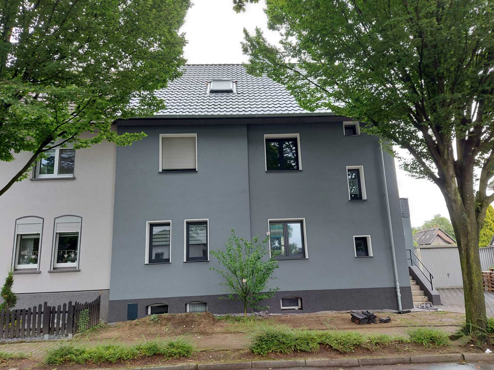 Graues Gebäude mit schwarz gerahmten Fenstern, neben einem helleren Gebäude, Bäume im Vordergrund.