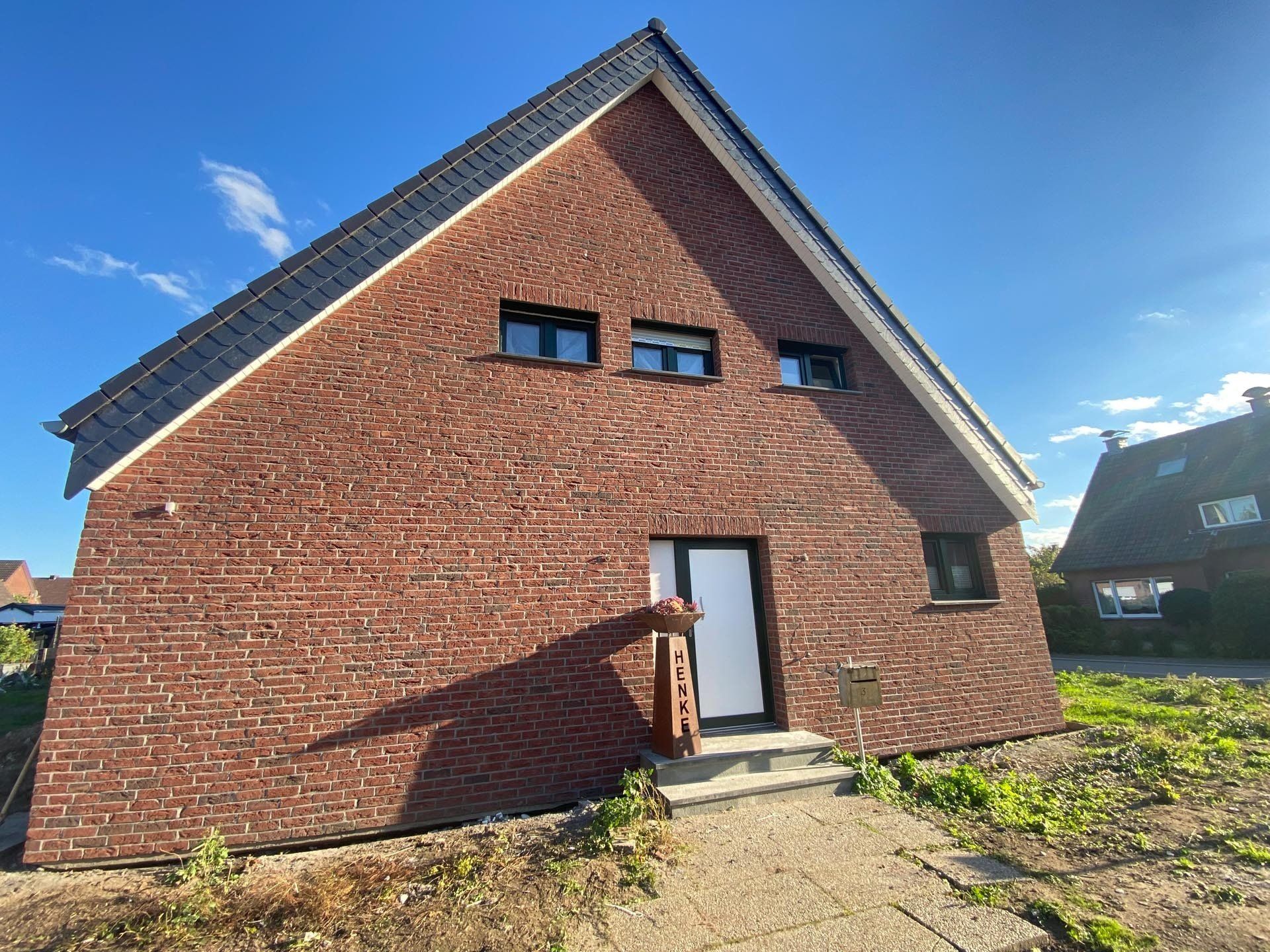 Backsteinhaus mit schwarzem Dach, Tür und Fenstern vor blauem Himmel.