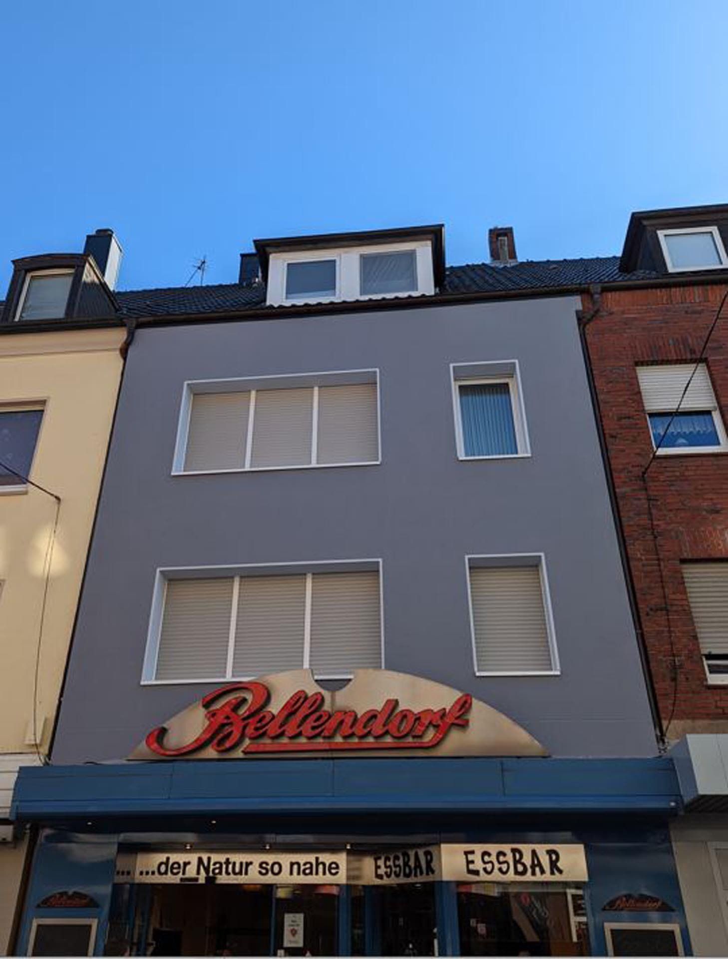 Gebäudefassade mit „Bellendorf“-Schild. Graue Außenfassade, blaue Markise. Rotes Schild, Himmel im Hintergrund.