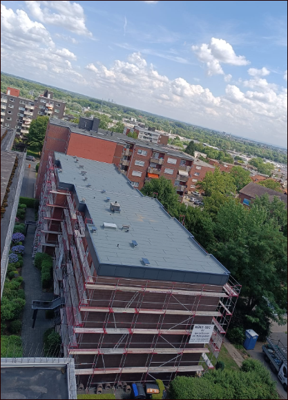 Eine erhöhte Ansicht eines mehrstöckigen Gebäudes mit Gerüst, umgeben von anderen Gebäuden und grünen Bäumen, blauer Himmel.