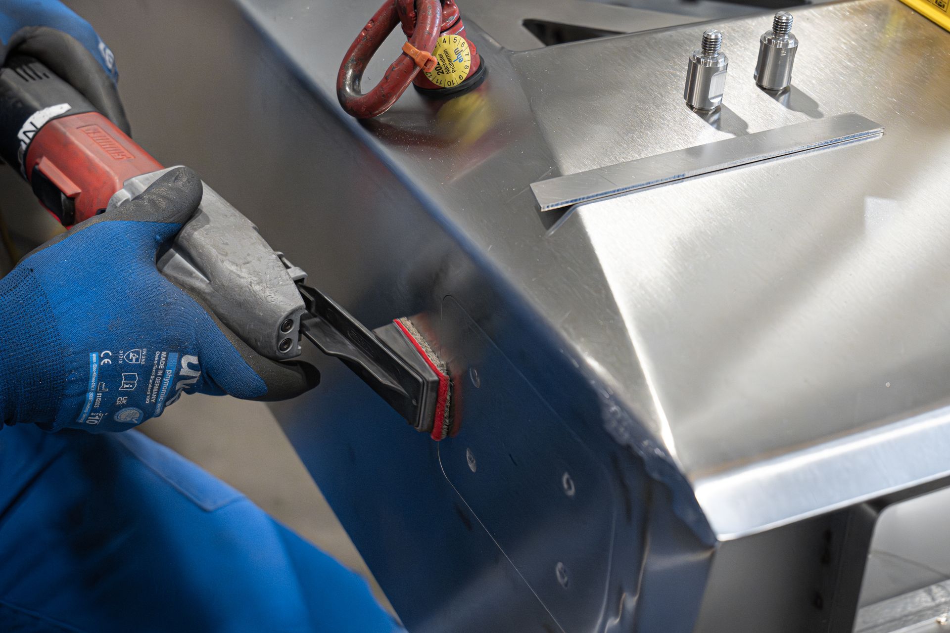 Ein Arbeiter mit Handschuhen schleift in einer Werkstatt mit einem Elektrowerkzeug ein Edelstahlbauteil.