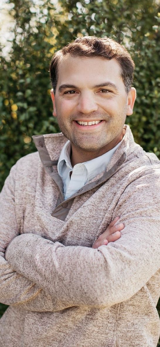Man smiling, arms crossed, wearing a light brown sweater with a button-down shirt in front of greenery.