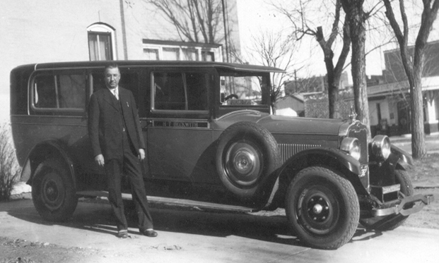 Man in suit stands beside a vintage sedan with spare tire; outdoors, trees, building in background.