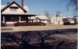 Tan house with porch, shrubs, and sidewalk. Other buildings and street in background.