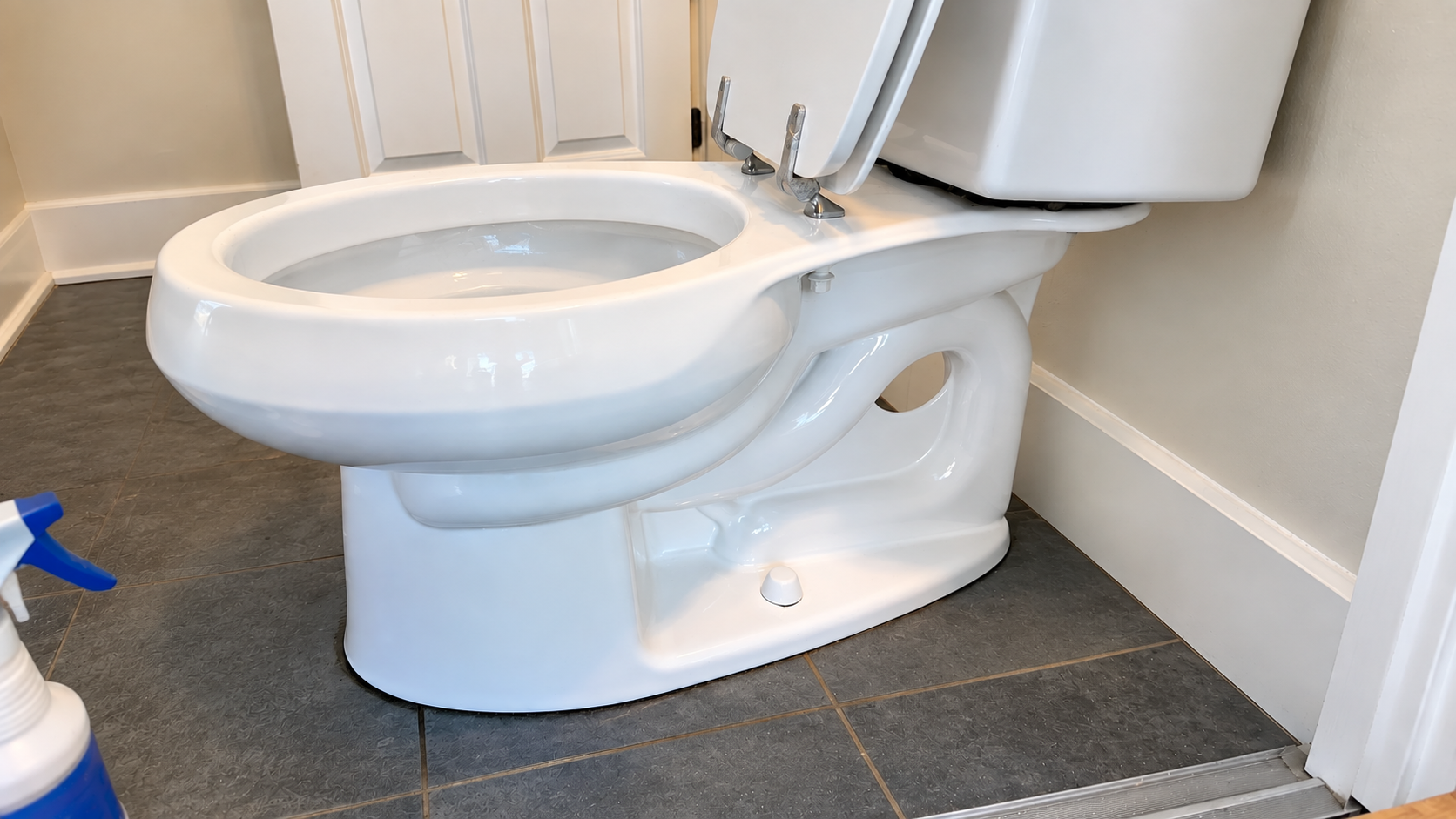 White toilet in a bathroom corner with the seat open and a blue spray bottle nearby on dark tile floor