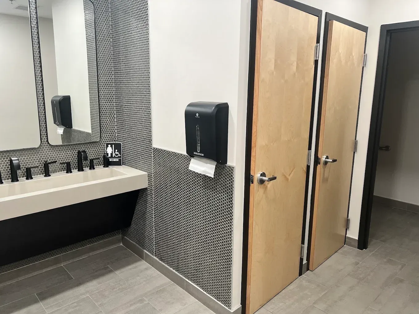 Public restroom with sinks, mirror, paper towel dispenser, and two stall doors on a gray tiled floor.