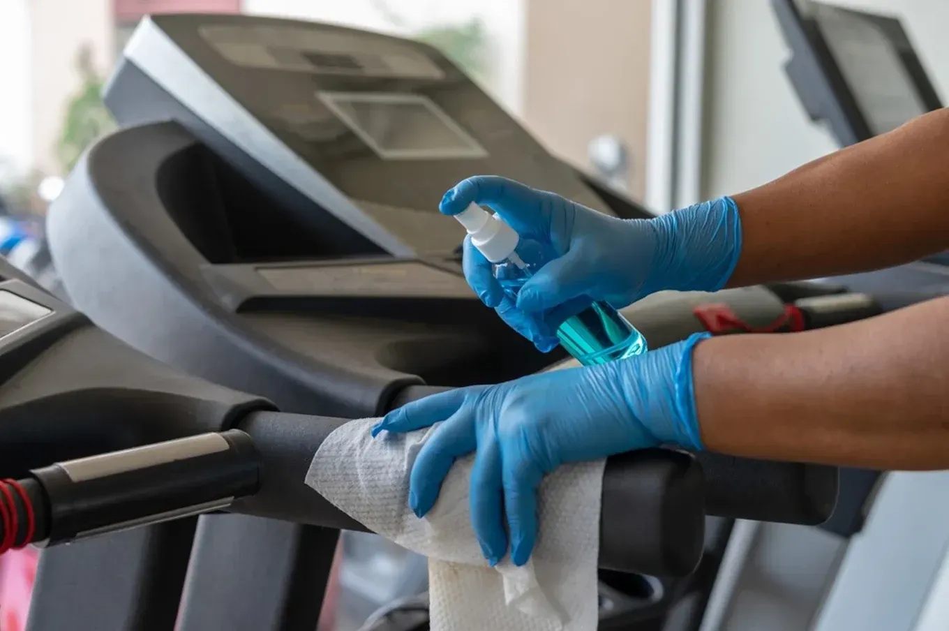 Gloved hands cleaning a treadmill console with a spray bottle and cloth