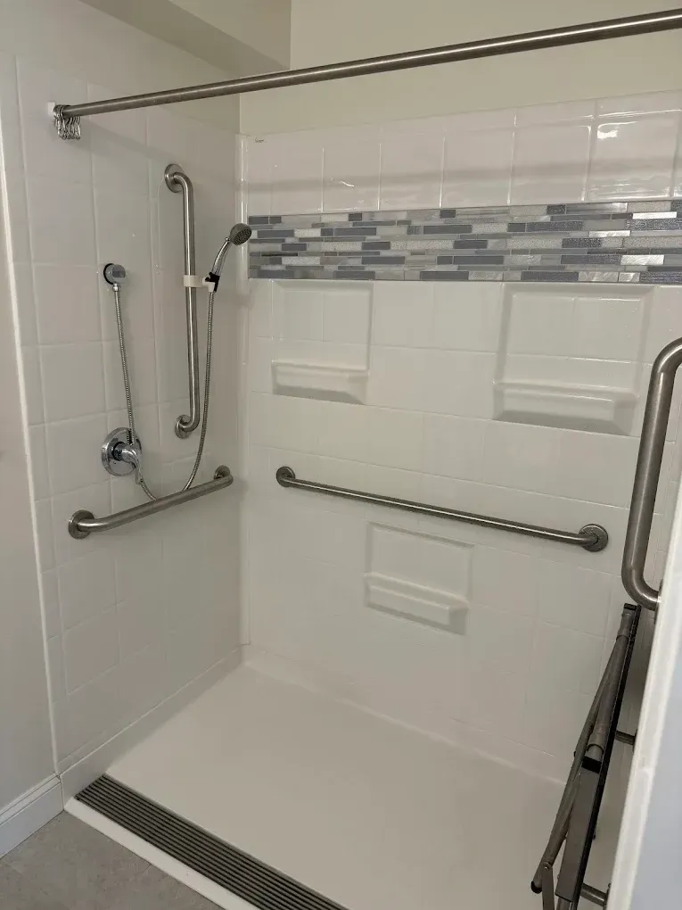 Accessible shower with grab bars, fold-down seat, handheld showerhead, and tiled walls.