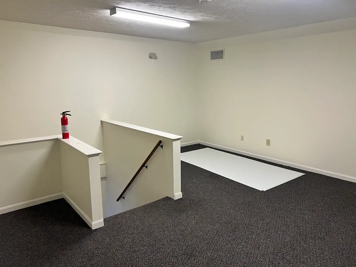 Empty carpeted room with a white half-wall, fire extinguisher, and overhead fluorescent lights