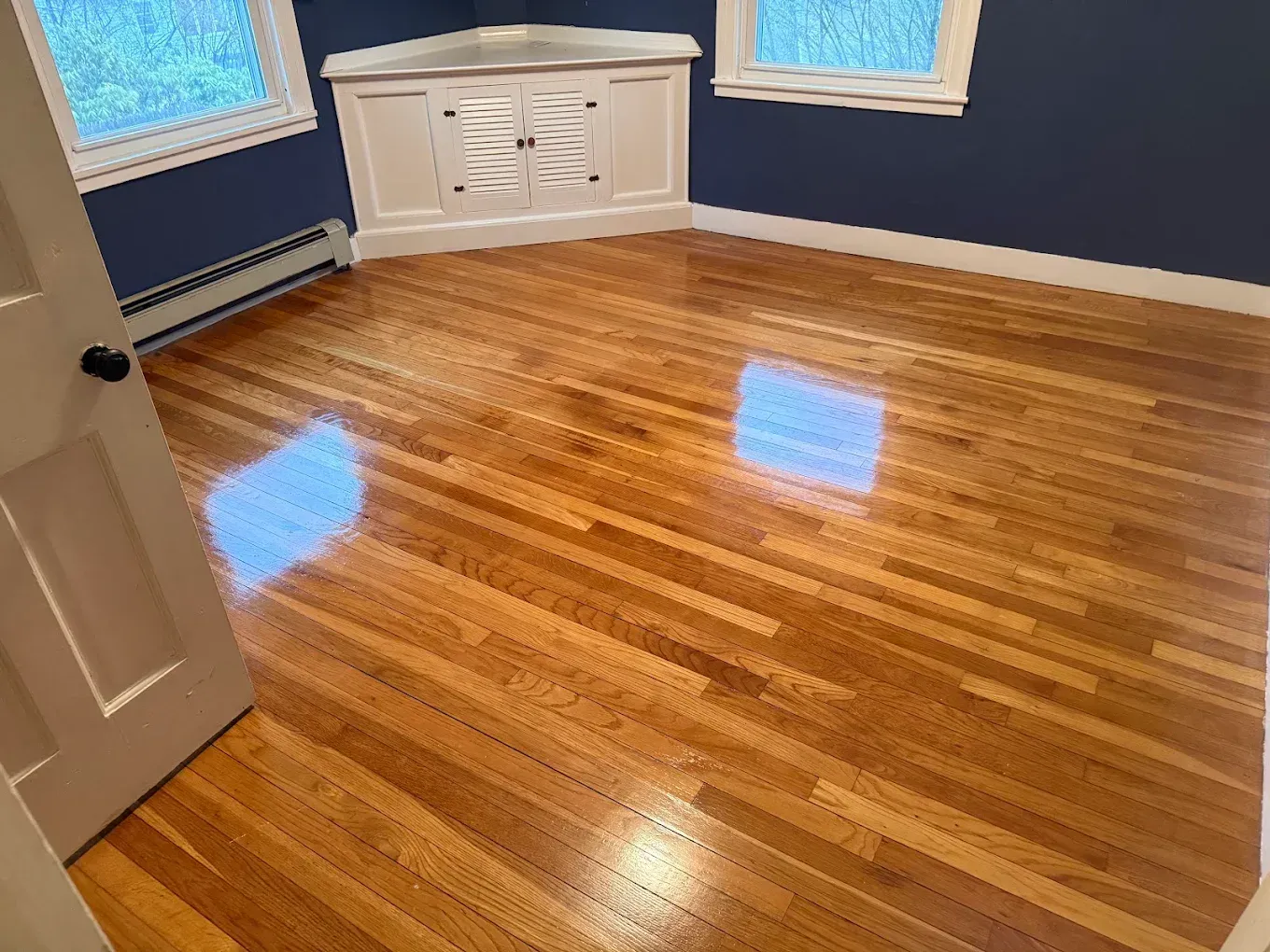 Empty room with polished hardwood floor, navy walls, and white corner cabinet under windows