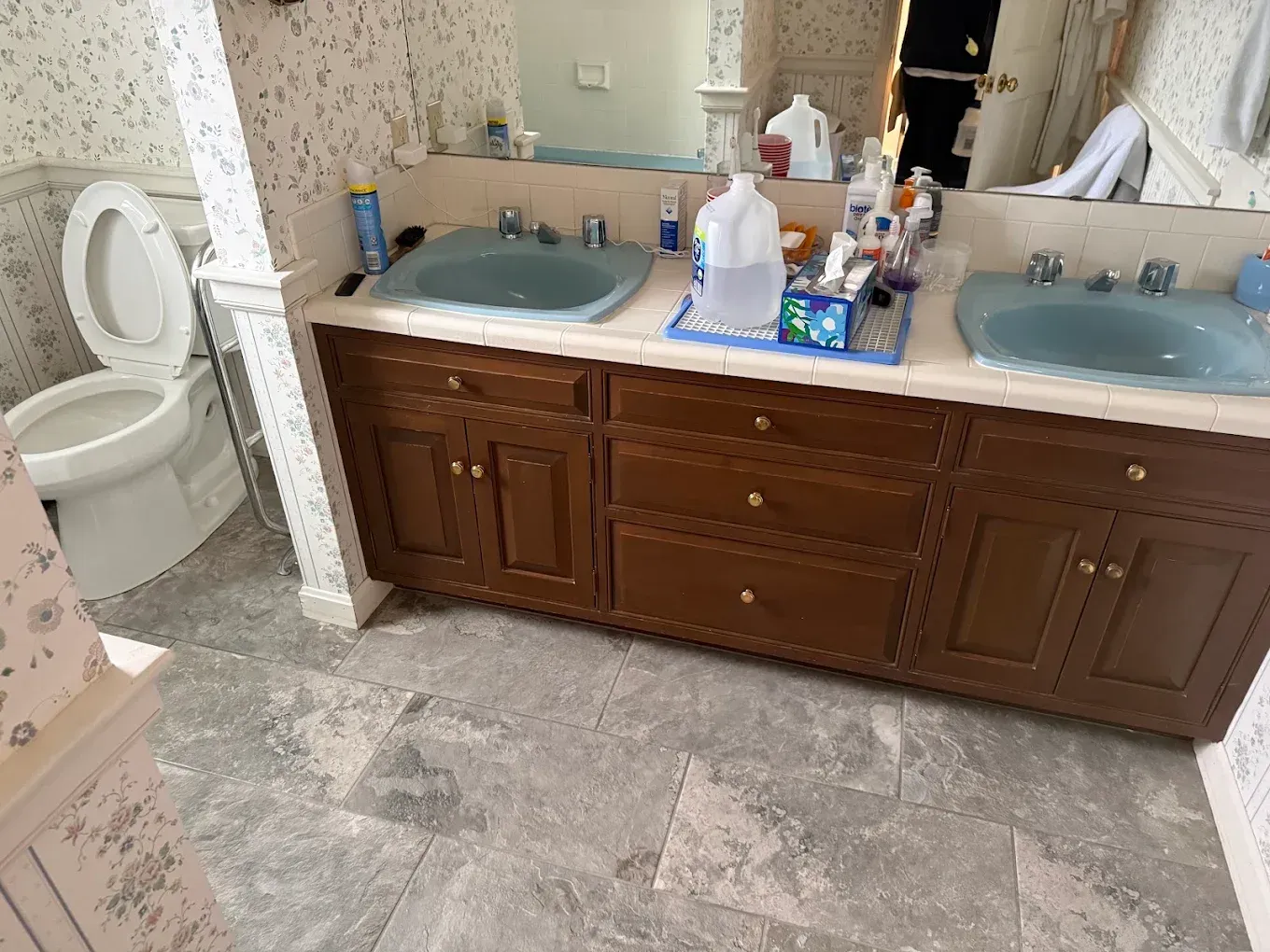 Bathroom with double sinks, wooden vanity, toilet, and tiled floor; toiletries and paper on the counter