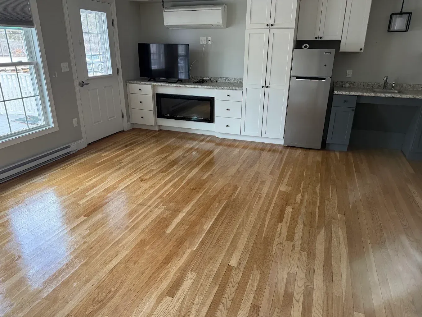 Empty living room with hardwood floors, white walls, fireplace, TV, and a refrigerator near the kitchen area