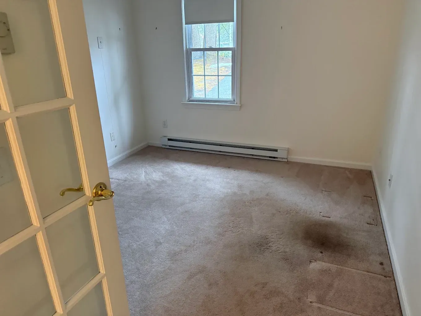 Empty beige bedroom with a window, white walls, and a stained carpet.
