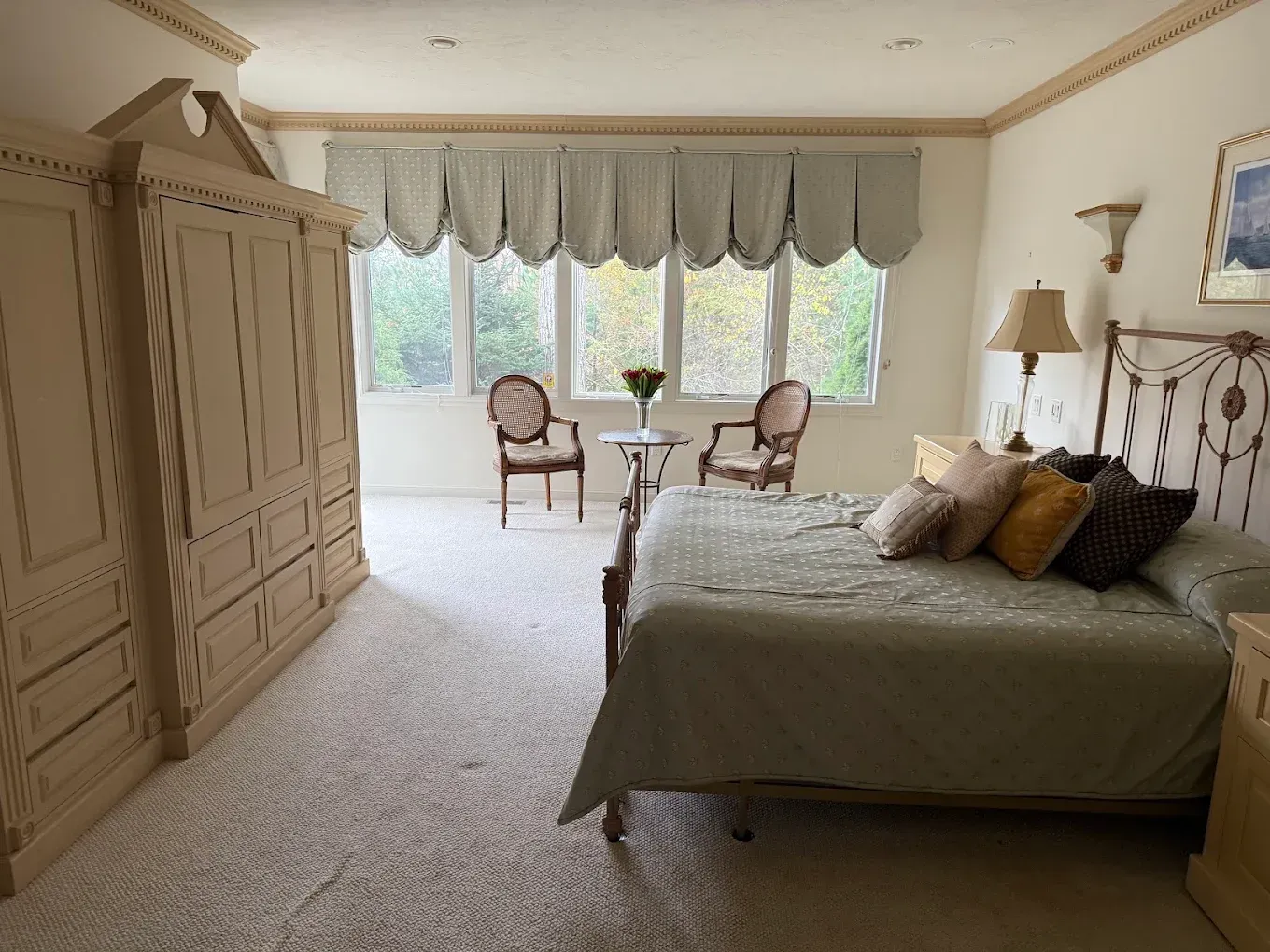 Bright bedroom with a large bed, armoire, and sitting area by a window with scalloped curtains.