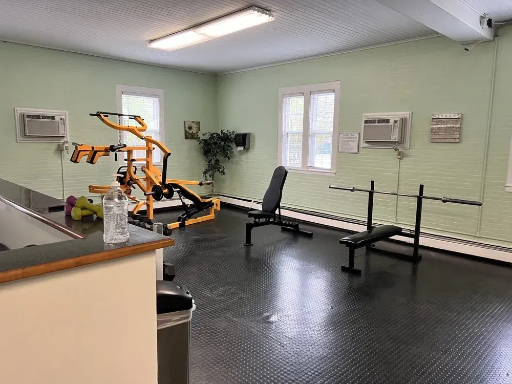 Small rehabilitation gym with exercise equipment, bench, wall-mounted air conditioners, and a bright windowed room.