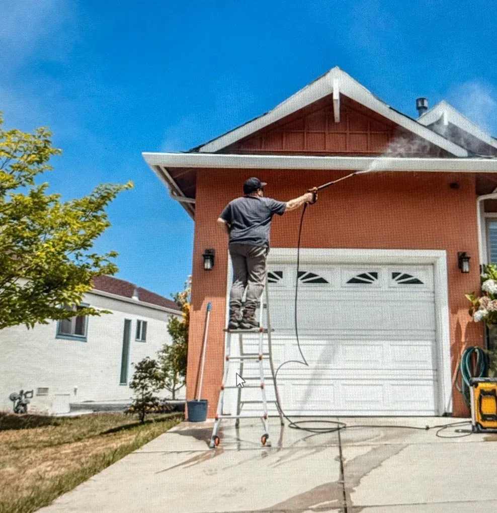 Worker on a ladder pressure-washing the roof of a red house on a sunny day