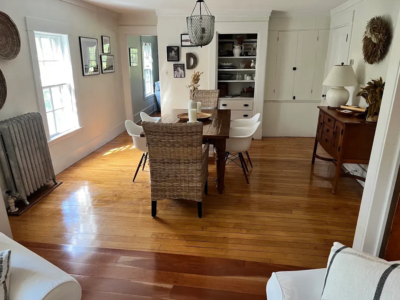 Bright dining room with wicker chairs, white table, wooden floors, and sunlight from tall windows.