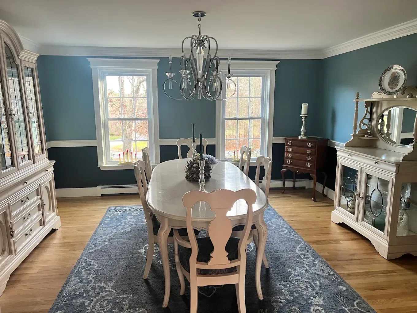 Formal dining room with teal walls, white furniture, chandelier, and a long table on a blue rug.