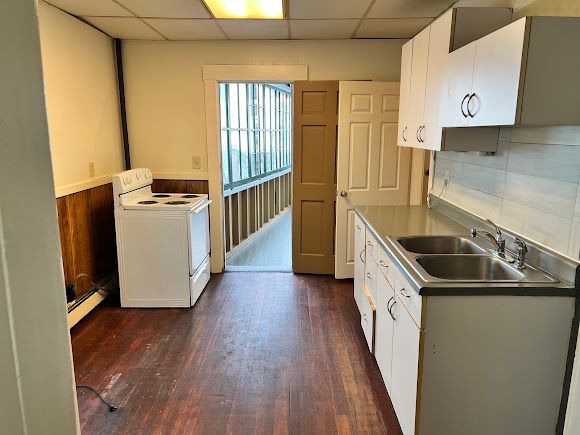 Narrow kitchen with white cabinets, stainless sink, stove, and a washer near a window.