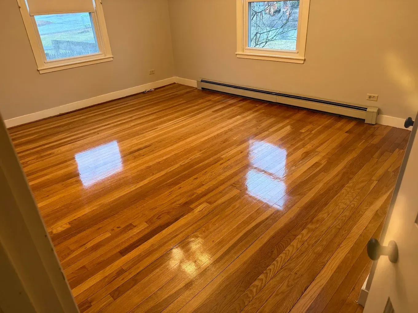 Empty room with polished hardwood floors, two windows, and beige walls.