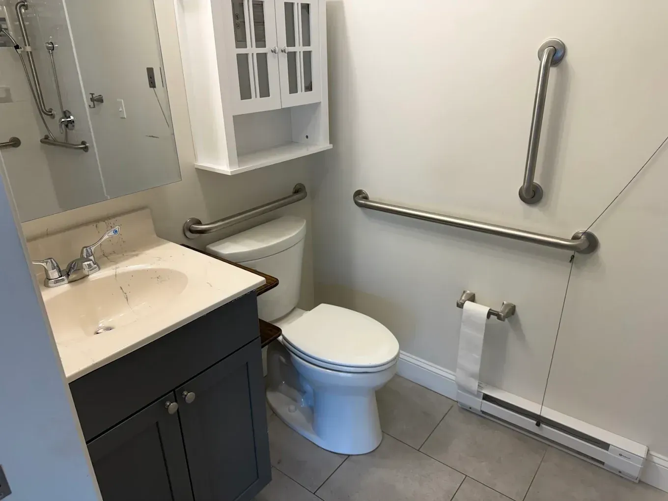 Accessible bathroom with toilet, sink, grab bars, and wall-mounted dispenser near a mirror.