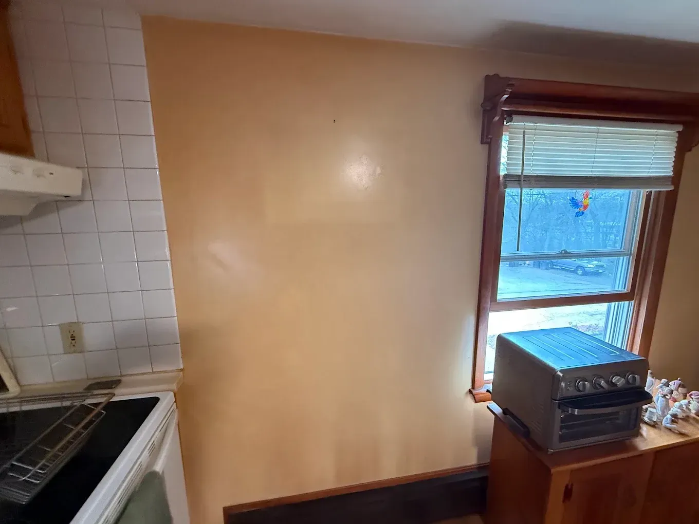 Small kitchen with tan walls, white tile backsplash, stove, and a window with blinds and a counter appliance.