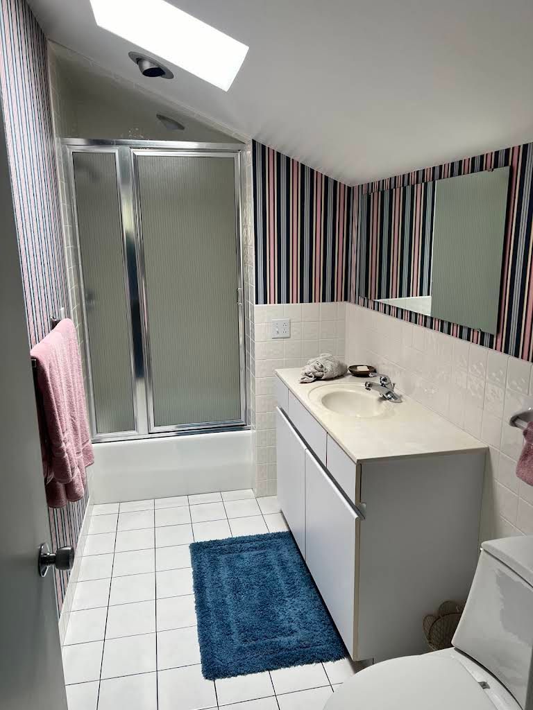 Small bathroom with shower, sink vanity, striped wallpaper, blue bath mat, and patterned tile floor.