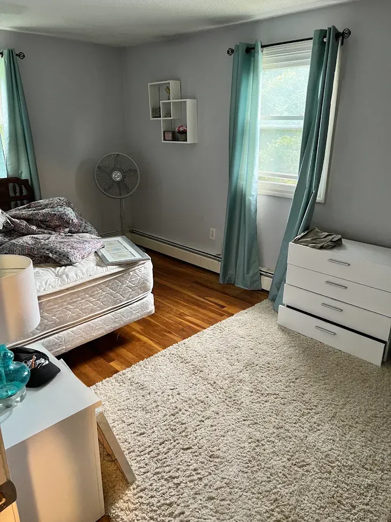 Bright bedroom with teal curtains, white dresser, bed, fan, and hardwood floor