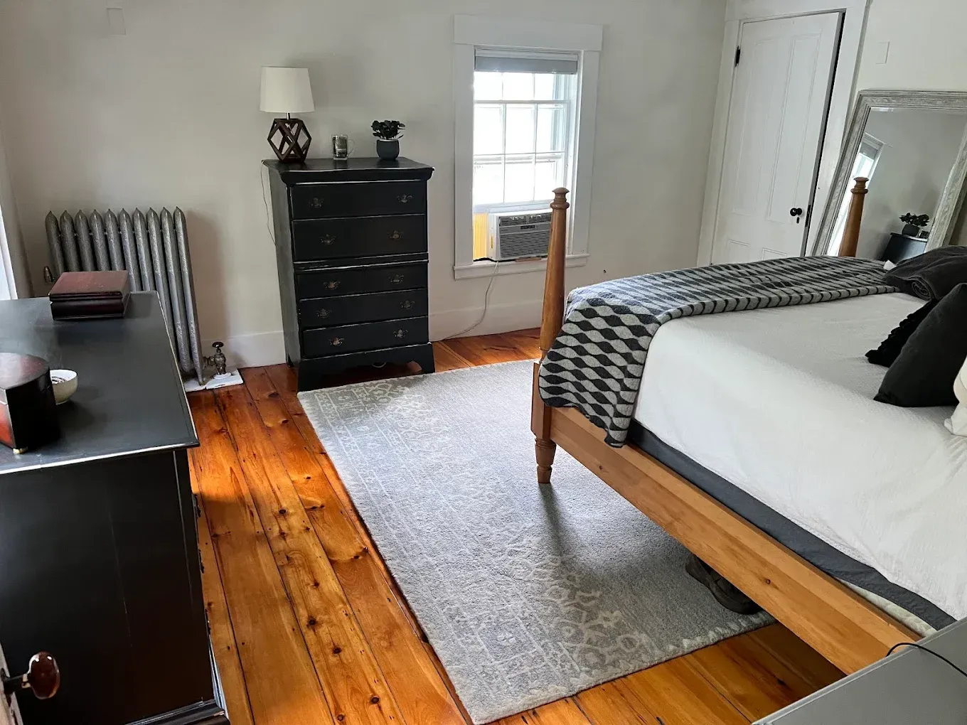 Bedroom with white bed, gray rug, dark dresser, desk, and exposed hardwood floor by a window