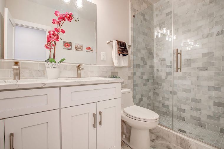White bathroom with vanity, mirror, toilet, and tiled shower, accented by a pink orchid.