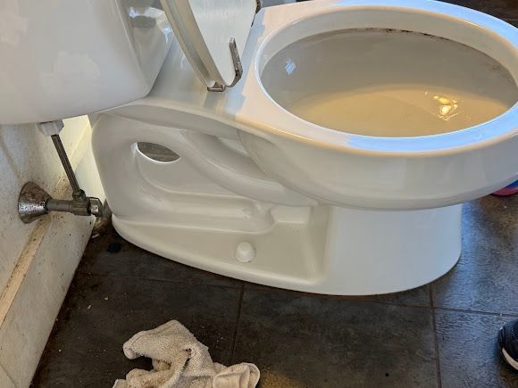 Bathroom with a toilet, exposed plumbing, and a cloth on the dark floor tiles.