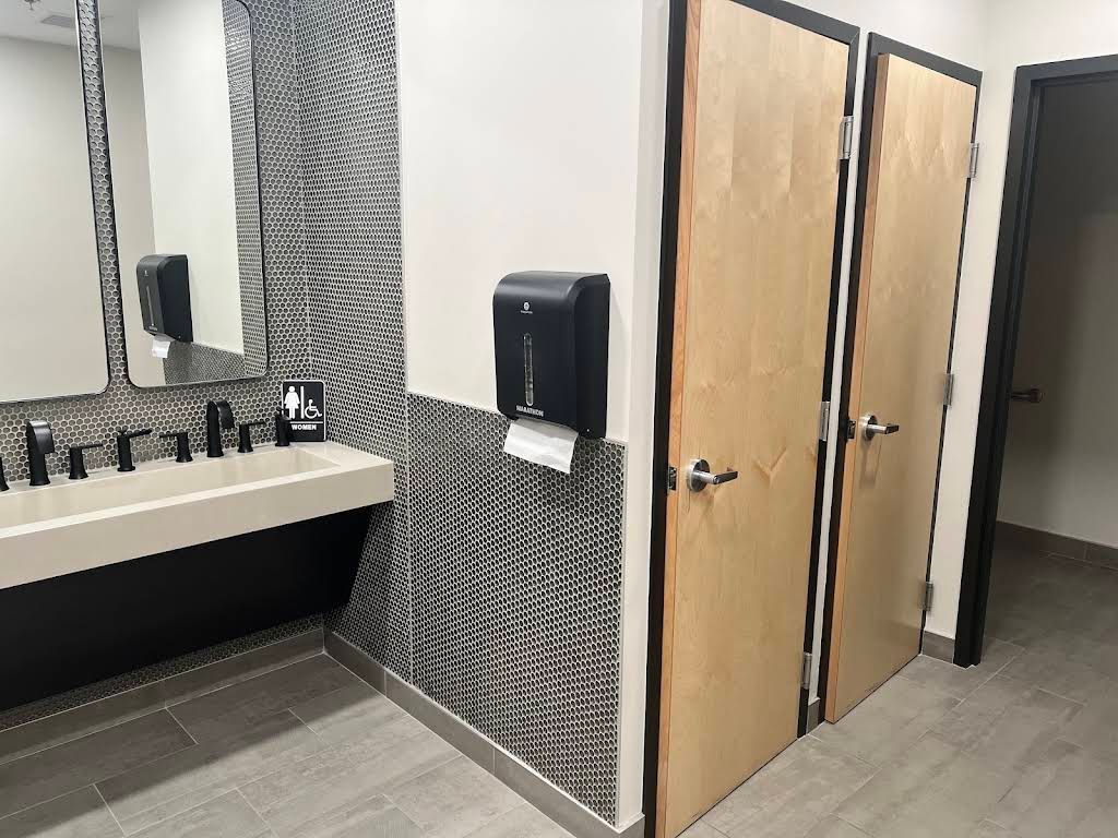 Public restroom with double sinks, large mirror, paper towel dispenser, and two wooden stall doors.
