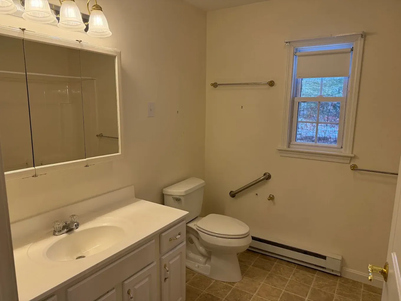 Small beige bathroom with vanity, mirror, toilet, grab bars, window, and baseboard heater.