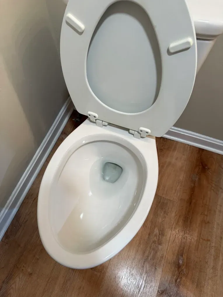 Open toilet with white seat and lid in a bathroom with wood-look floor and beige wall.