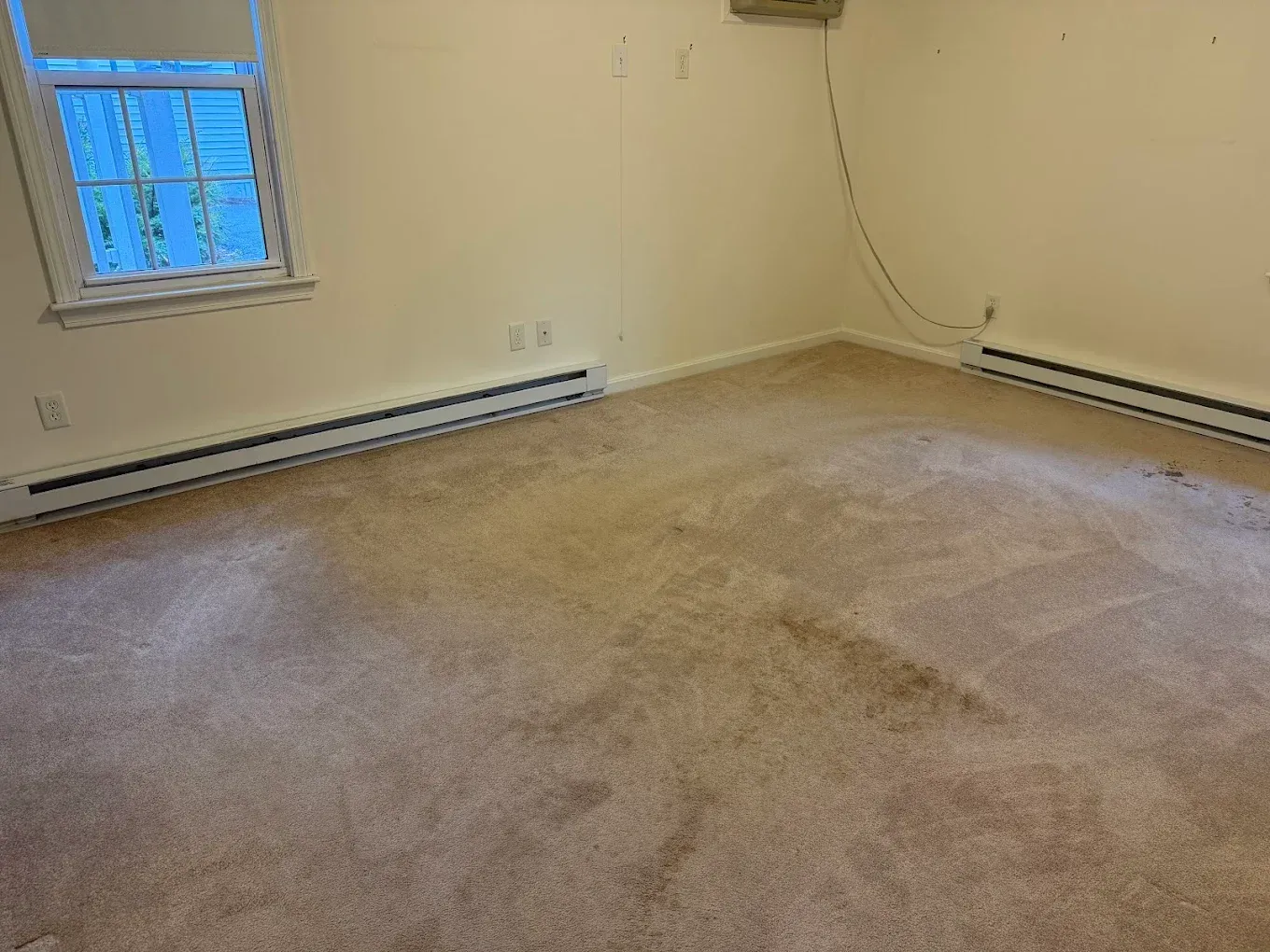 Empty beige room with stained carpet, small window, and baseboard heaters along the walls
