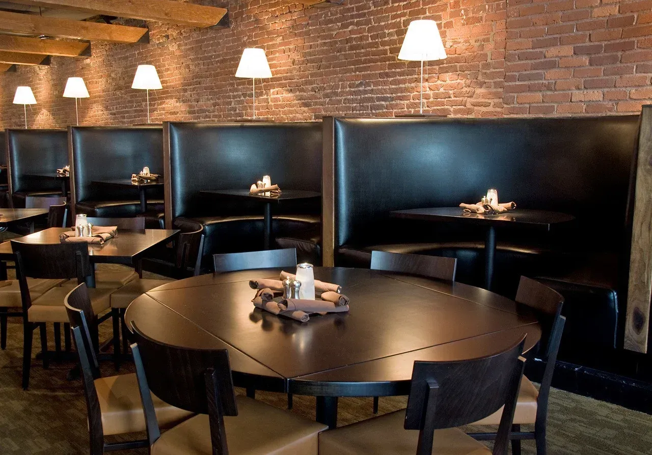 Cozy restaurant booth area with round tables, black chairs, and brick walls lit by hanging lamps