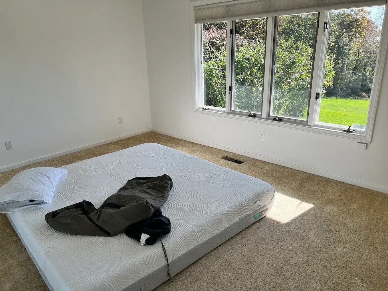 Empty bedroom with a mattress and folded coat on the floor, sunlight through large windows