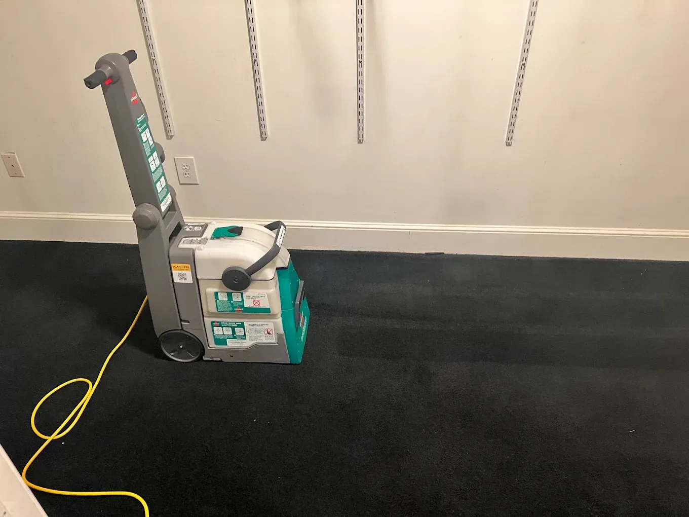 Portable carpet cleaning machine on black carpet beside a wall, with yellow cord attached.