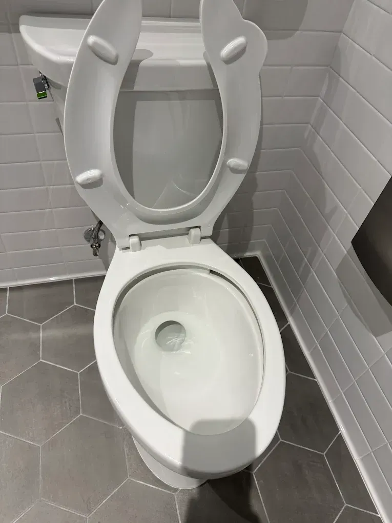 Open toilet in a small gray-tiled bathroom with a white seat and lid up